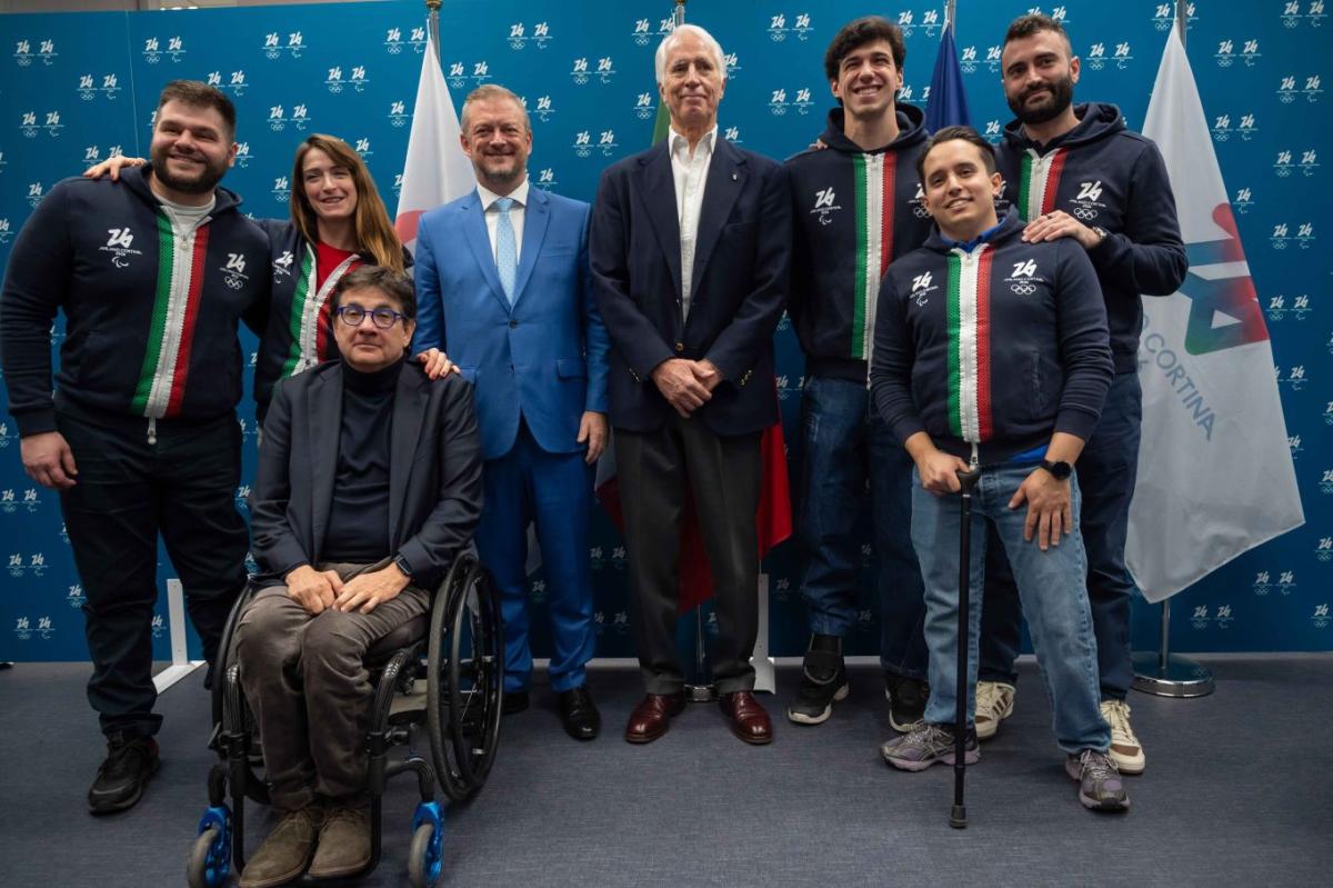 Eight people, including Paralympic athletes and officials, pose for a photograph in front of a blue banner with logos of the Milano Cortina 2026 Games