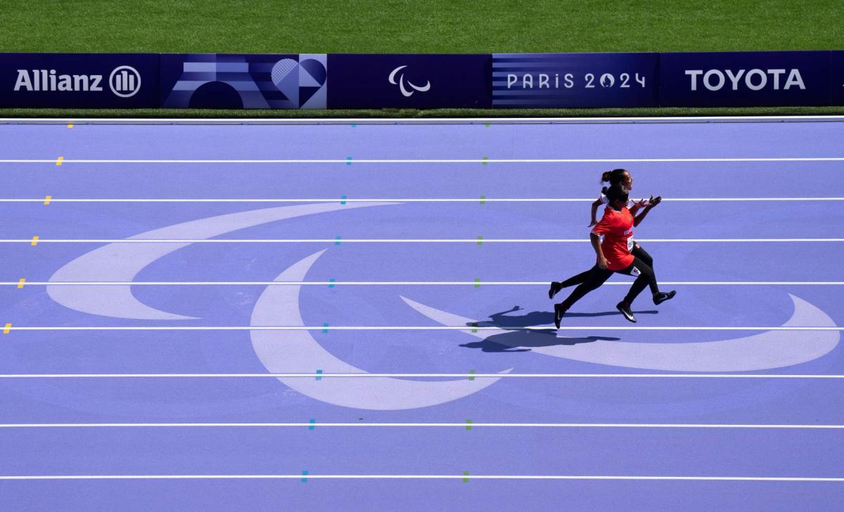 The purple track of the Paris 2024 Paralympics. There is a female athlete competing with her sighted guide
