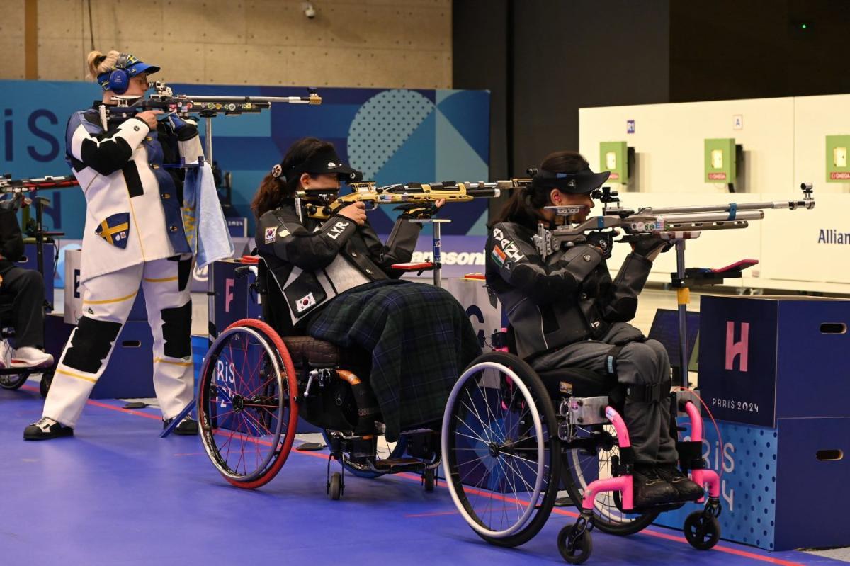A standing woman and two women in wheelchairs competing in a rifle shooting range