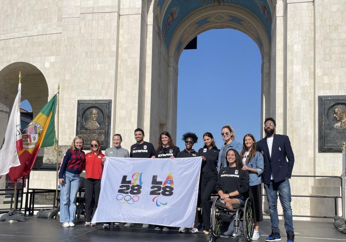 11 people including one female Paralympian pose for a group photo in front of a stadium. They are holding a white flag with the logos of the LA Games