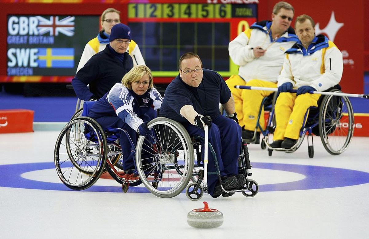 A male wheelchair curling athlete is releasing a stone with a stick. A female player is holding his wheelchair, while four athletes are watching him on the ice.