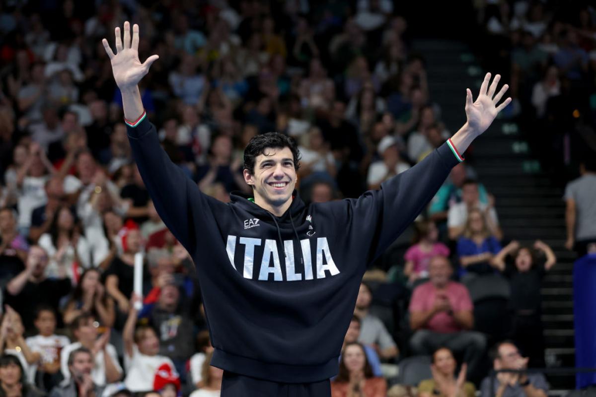 A male Para swimmer wearing a hoodie that says Italia raises his arms in front of a crowd