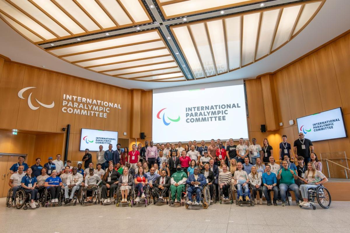 A group photo taken during the IPC Athletes' Forum - more than 70 athletes and officials are smiling in front of a screen with the IPC logo