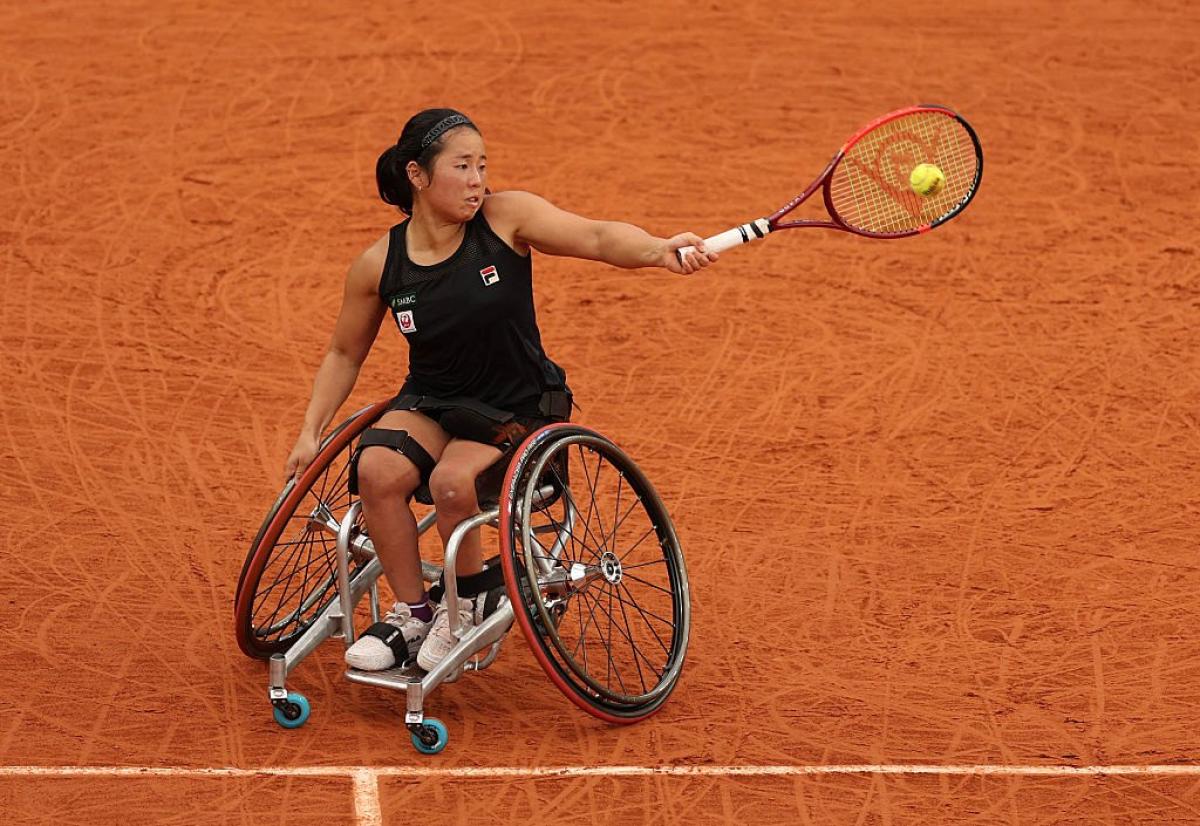 A female wheelchair tennis player plays a forehand on red clay court