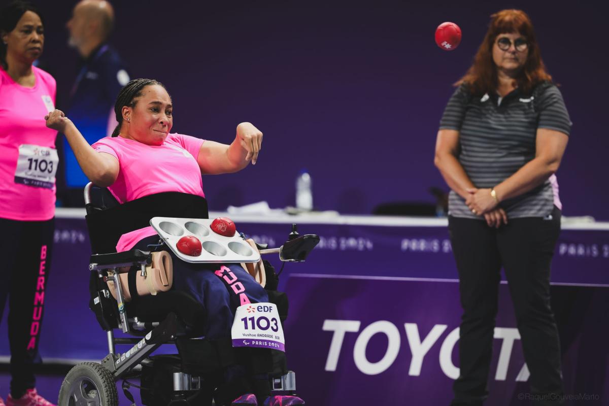 A female boccia athlete wearing a pink uniform is throwing a red boccia ball