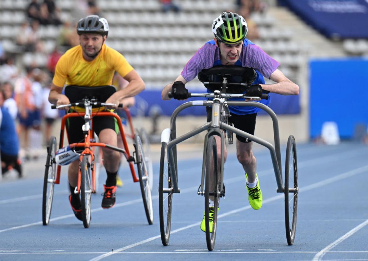 Two athletes are racing on a blue athletics track. They are running using a metal frame with three wheels