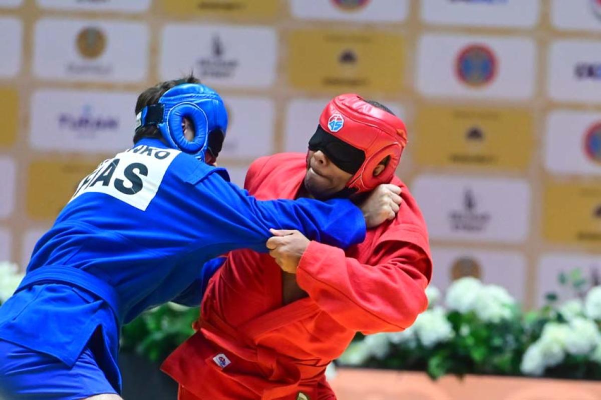 Two male athletes wearing blindfolds are wrestling - one athlete is wearing a red uniform and helmet while his opponent is wearing a blue helmet and gear