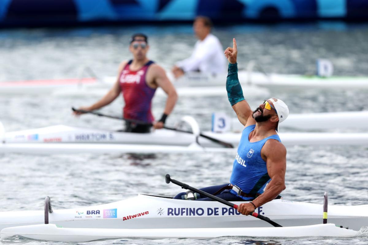 A male Para canoe athlete celebrates after winning his race - he is pointing his right index finger to the sky.
