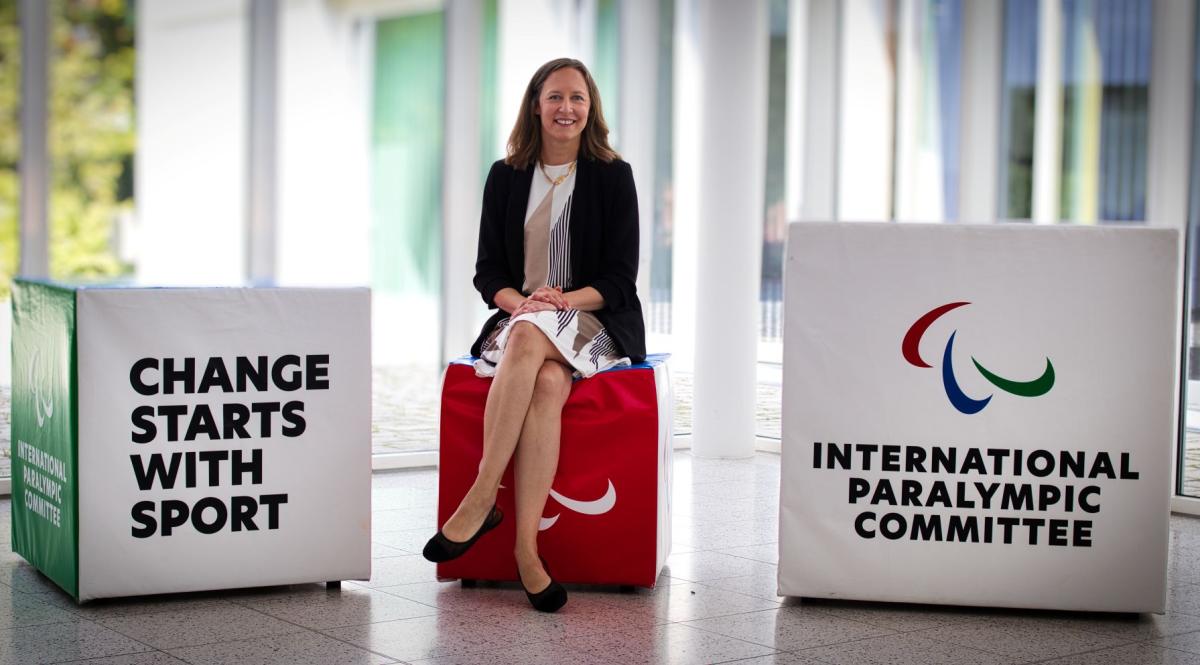 IPC Deputy CEO Kristina Molloy sitting on a red block in the IPC headquarters in Bonn, Germany. There are two blocks next to her - one with the IPC logo and motto "Change Starts with Sport"