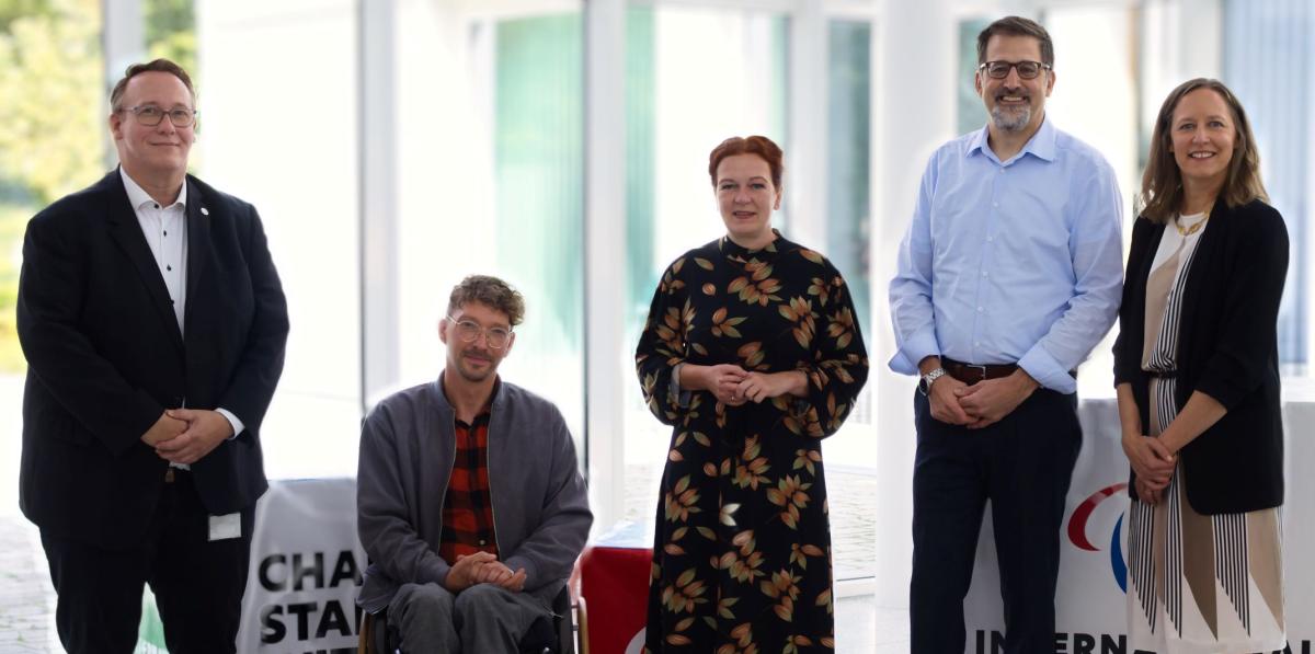 Five people pose for a photo in front of glass windows at the IPC headquarters in Bonn, Germany