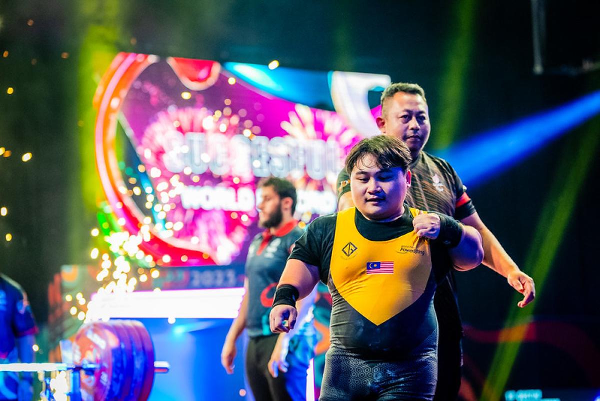 A short stature Para powerlifter on stage with three people in the background