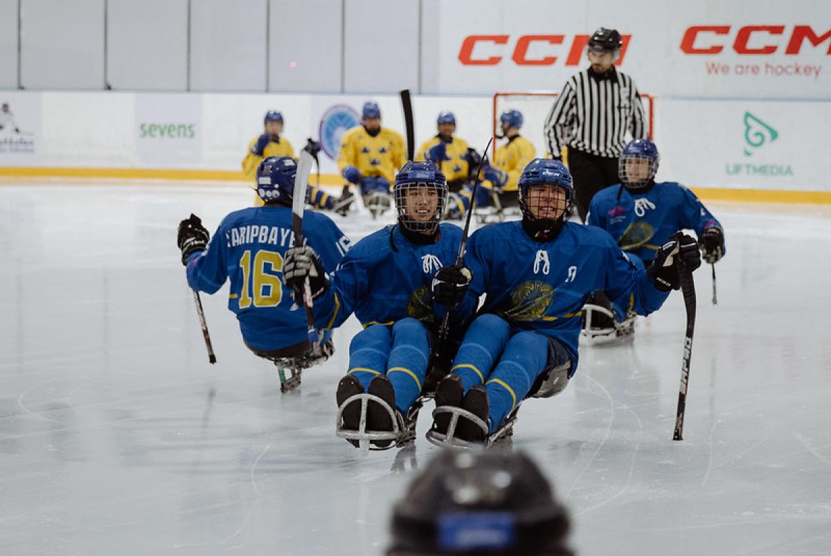 A group of four Kazak Para ice hockey players on ice with an official and four other players in the background