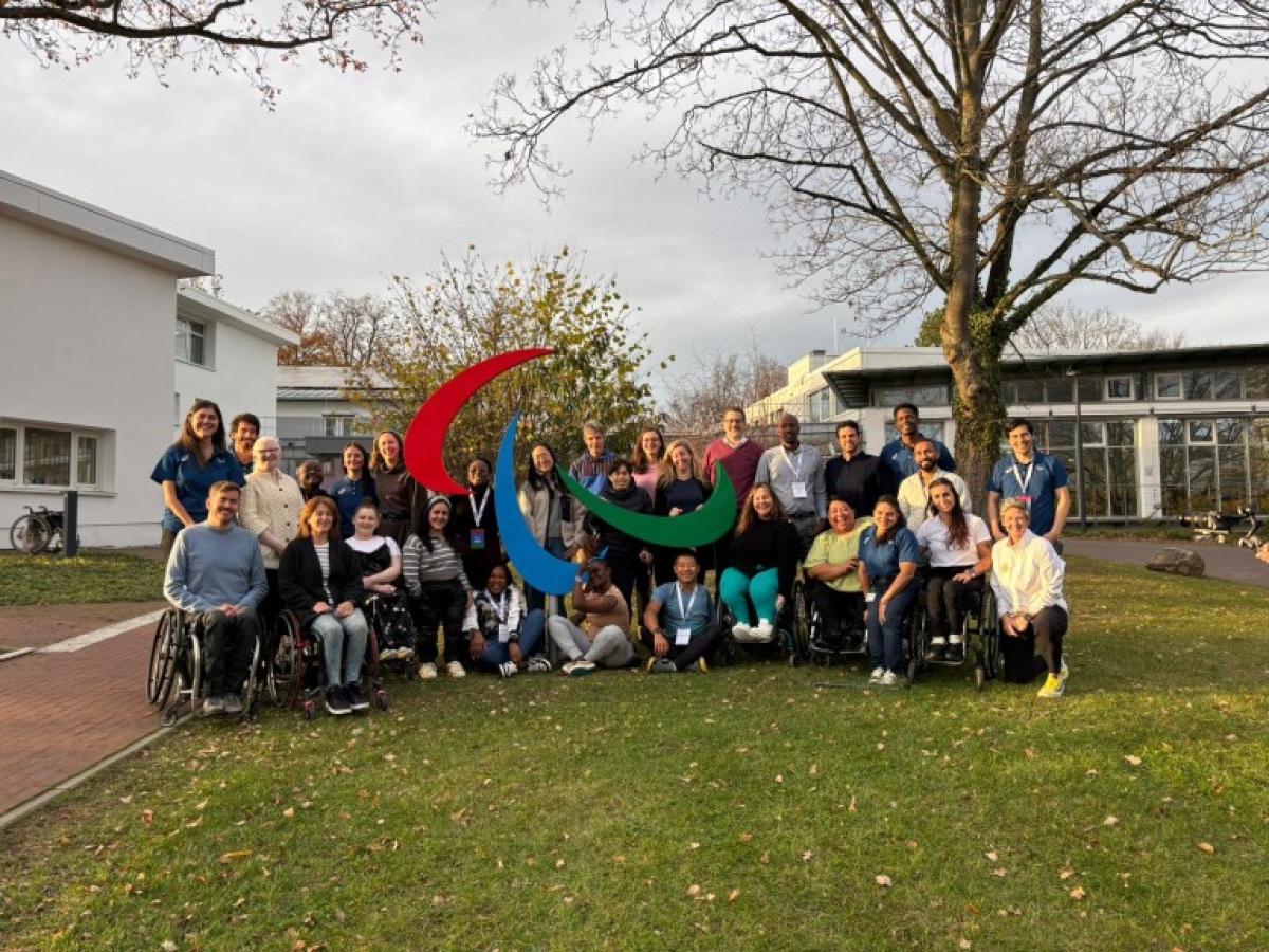 Twenty retired Para athletes take a photo in front of the Agitos symbol at the IPC Campus