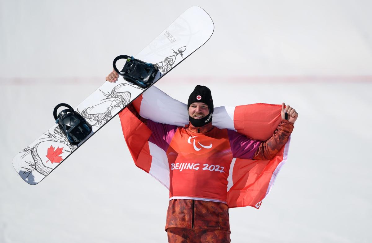 A male Para snowboarding athlete holds a snowboard and celebrates