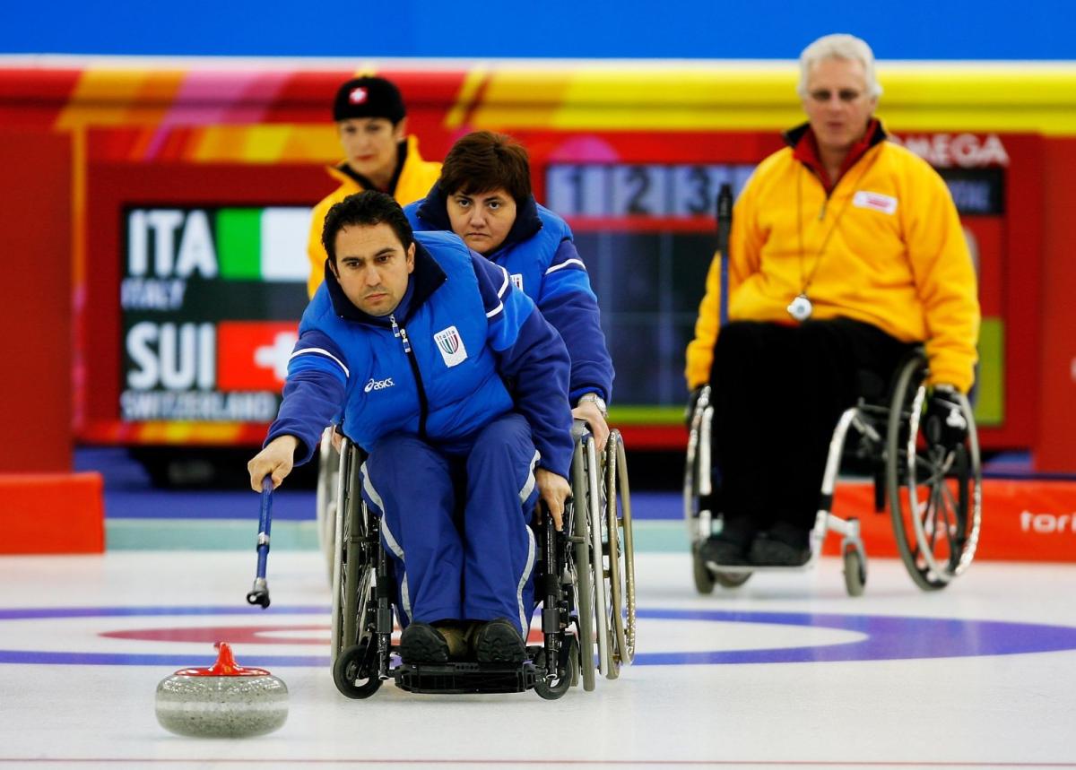 A male wheelchair curling athlete is delivering a stone with a stick during competition at Torino 2006.