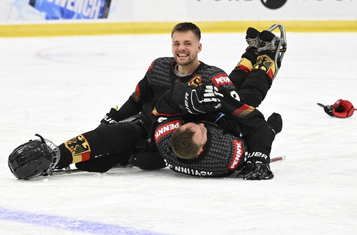 A male Para ice hockey player smiles on the rink, celebrating with his teammate who is lying on the ice