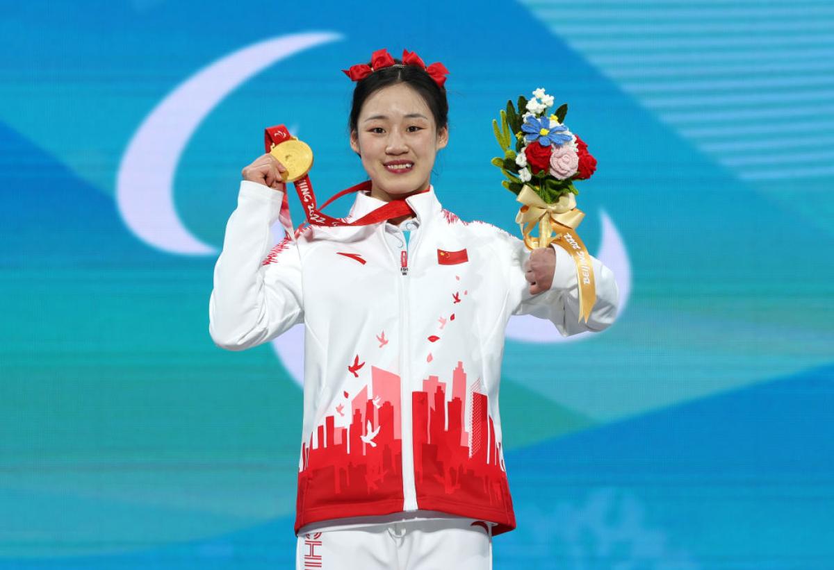 A female athlete with red ribbons on her head smiles on the podium while holding a flower bouquet and a gold medal