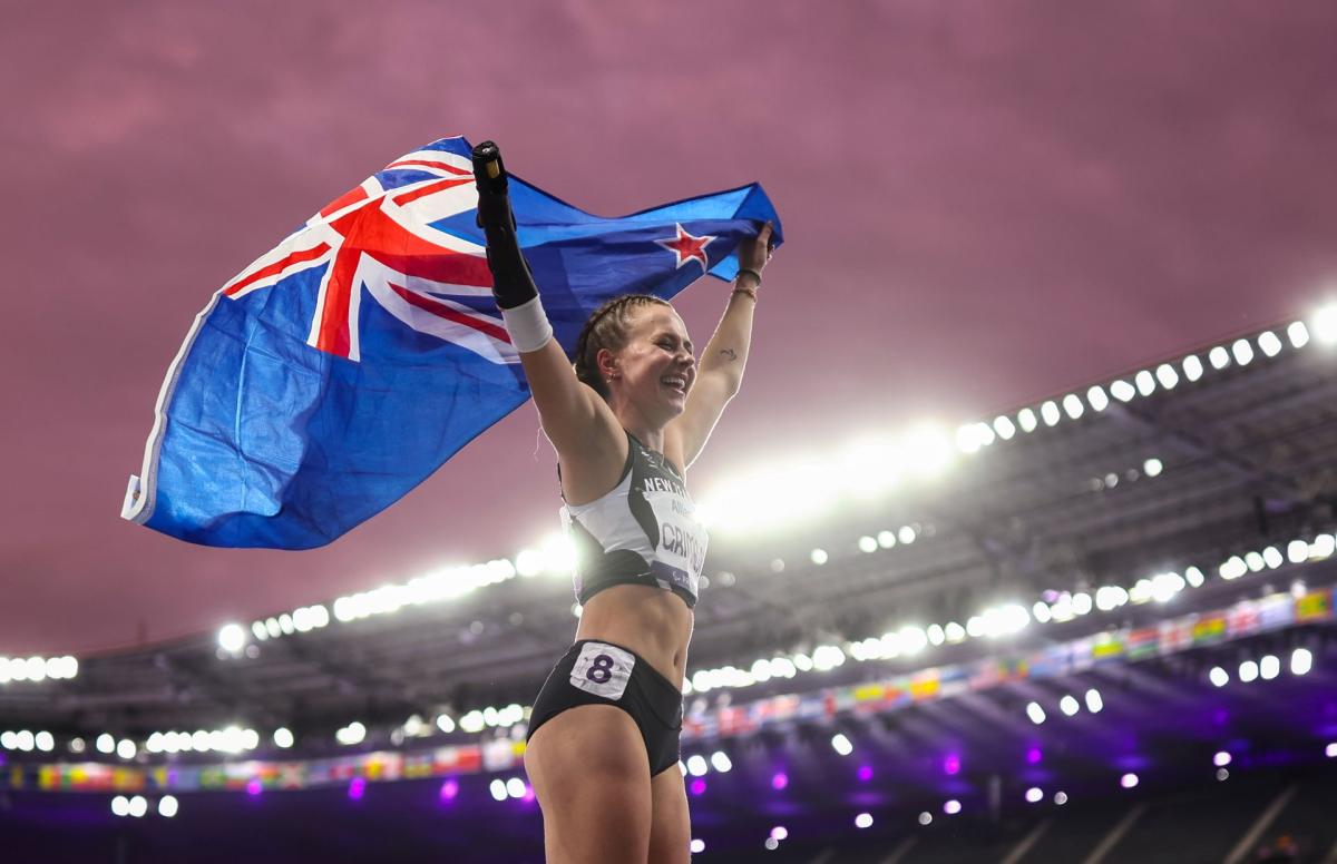 A female Para athlete wearing a track suit is holding New Zealand's flag in an outdoor stadium