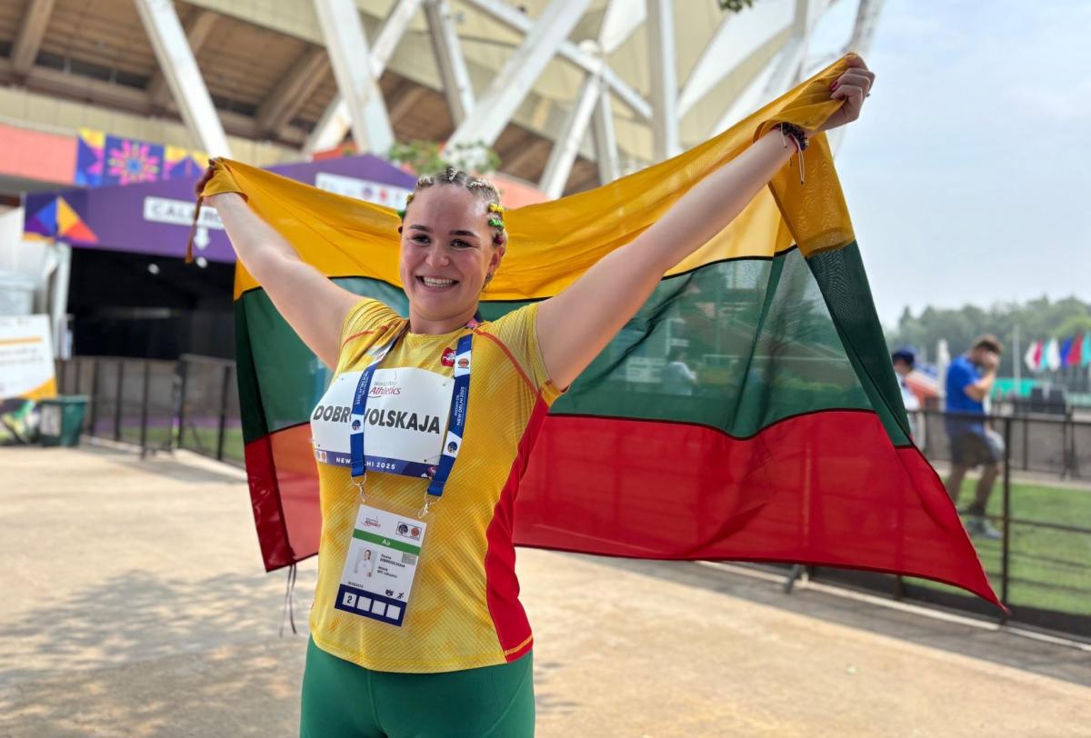 A female athlete wearing a green, yellow and red athletic gear is holding the Lithuanian flag in front of a stadium