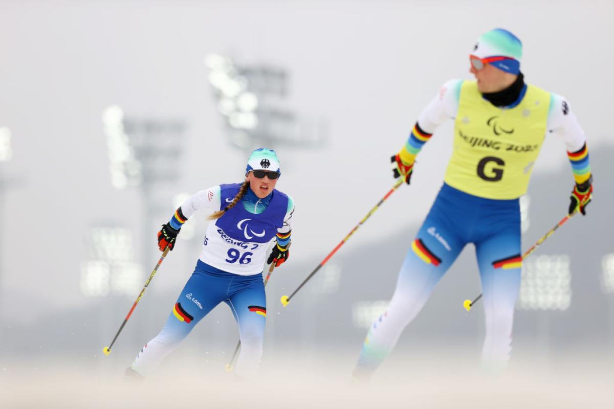 A female skier is competing behind a sighted male guide, who is looking back at her