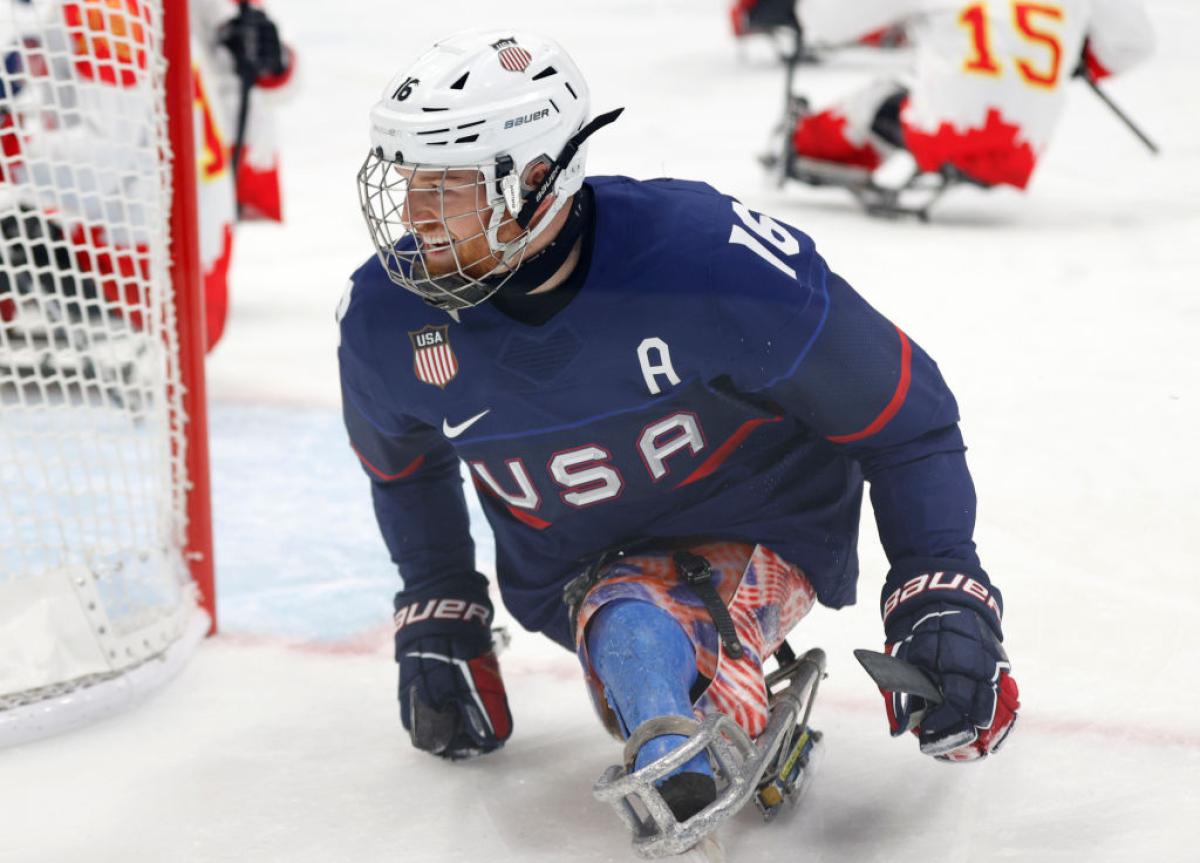 A male Team USA Para ice hockey player smiles and skates past the other team's net