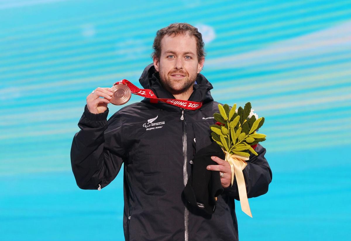 A male Para alpine skier celebrating his win, by holding a bronze medal and flowers