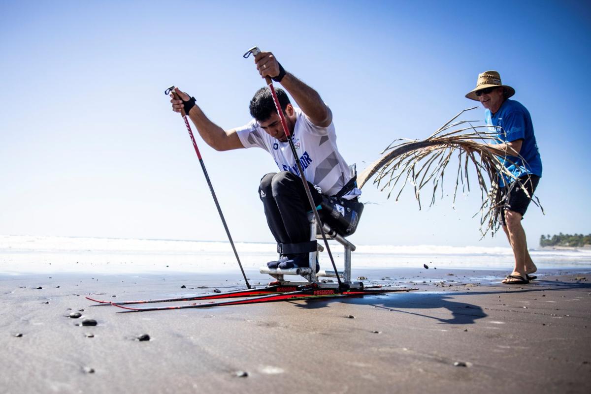 A male Para athlete is training on sit-skis on the beach, propelling forward using two poles.