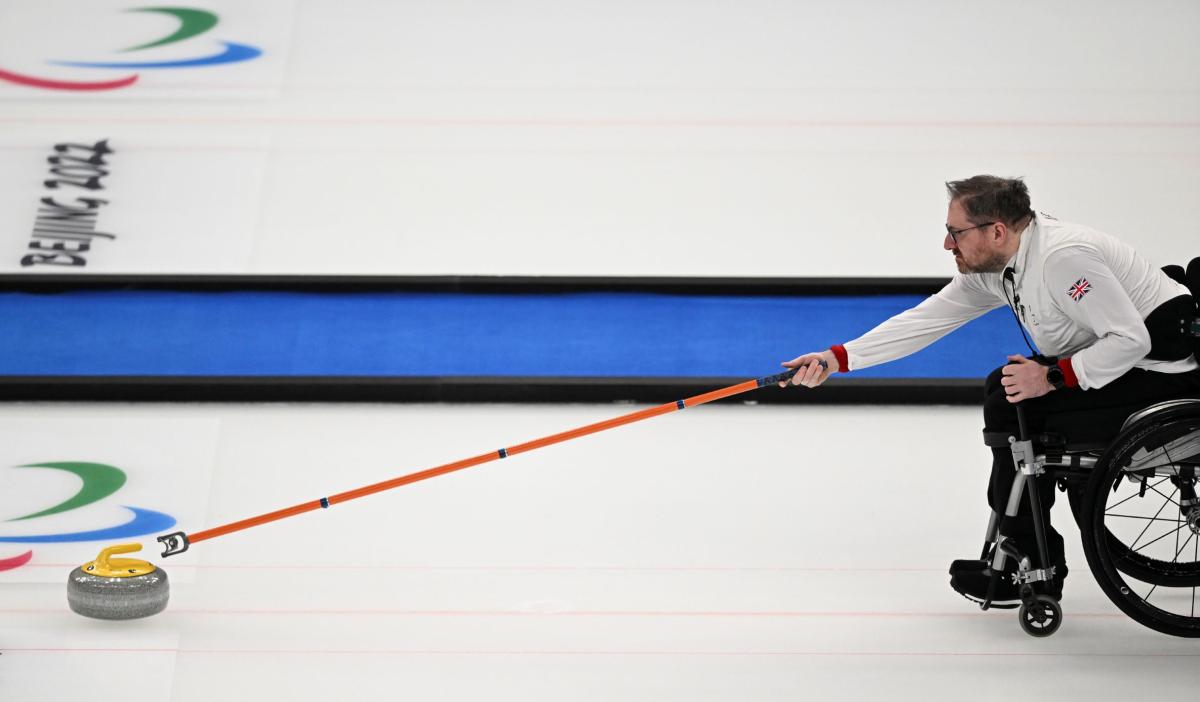 A male wheelchair curler, releases a curling stone on the ice using a delivery stick