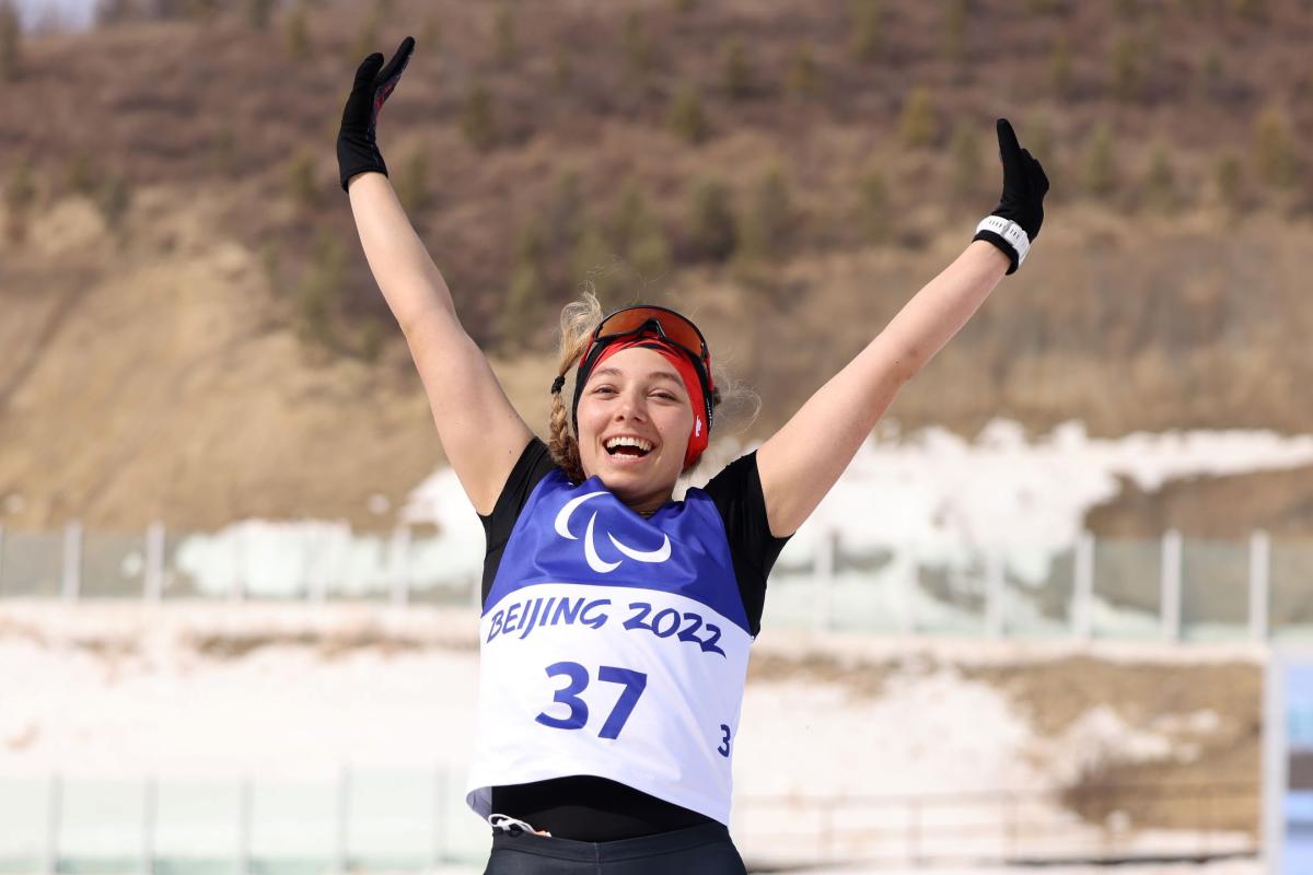  A female Para biathlon athlete,  jumping with her arms in the sky, while smiling at the camera