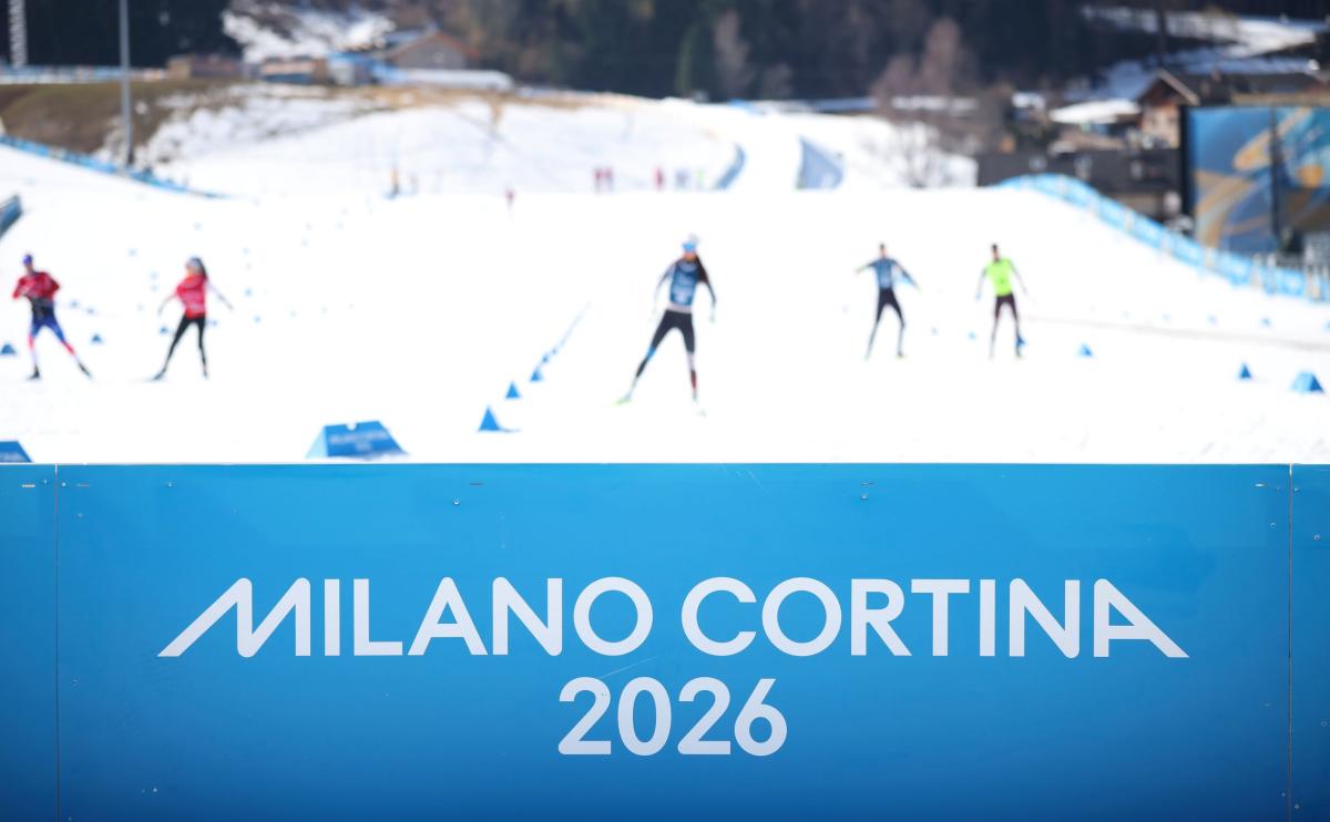 A banner with Milano Cortina 2026 written on it, while in the back para biathlon athletes are training