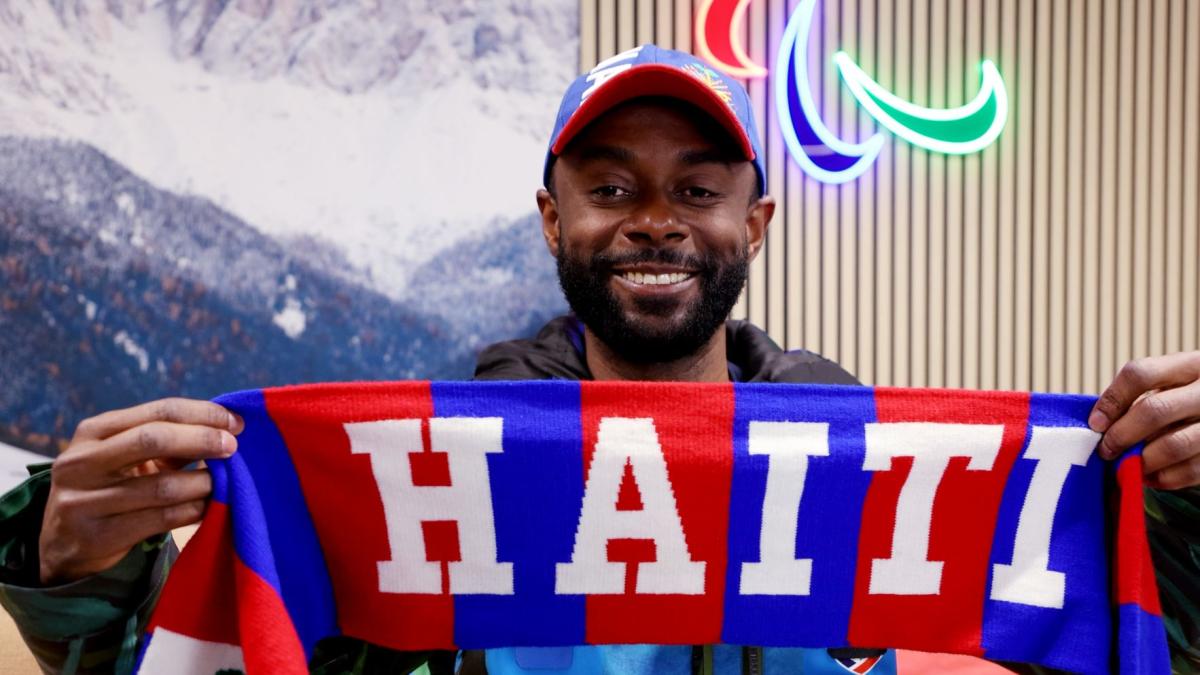 A male athlete wearing a cap holds a blue, red and white towel that says "Haiti" 