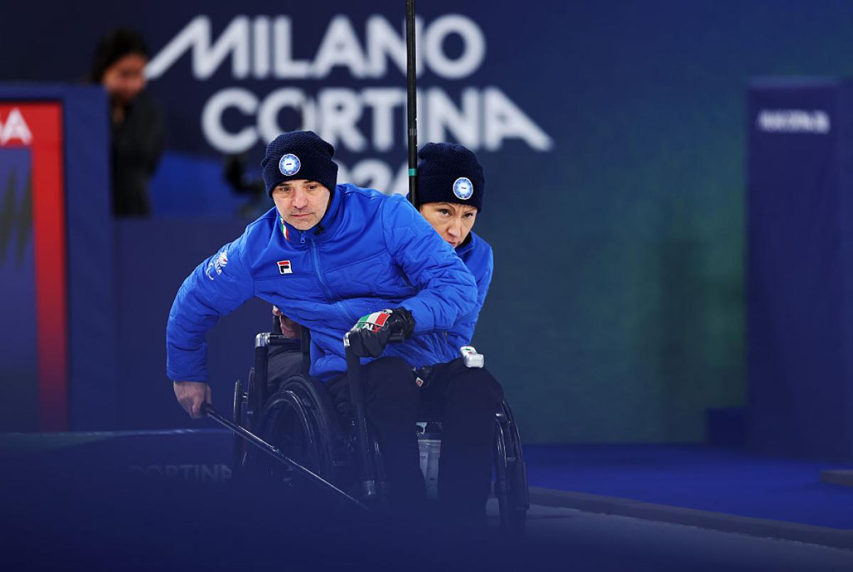 Two wheelchair curlers wearing blue uniforms are competing in front of a banner that says Milano Cortina 2026