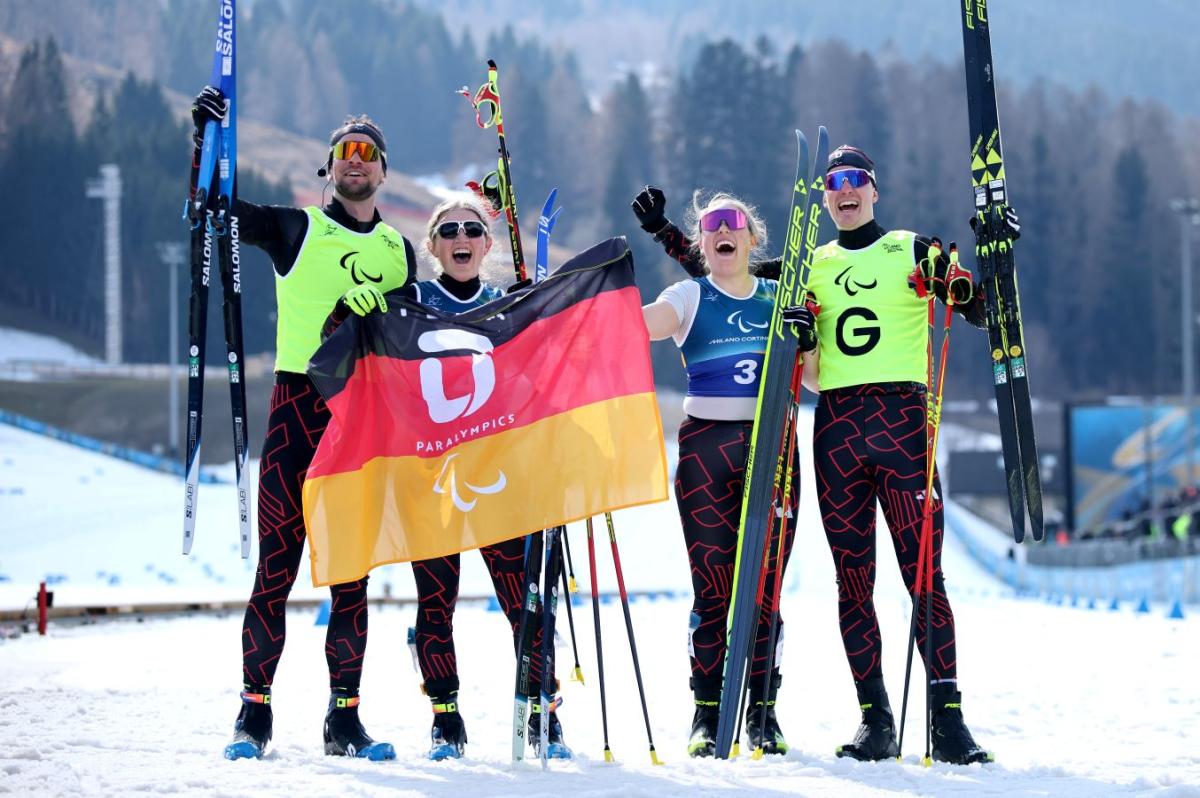 Two female Para cross-country skiers celebrating after competition waving their skis and the German flag, alongside their male guides