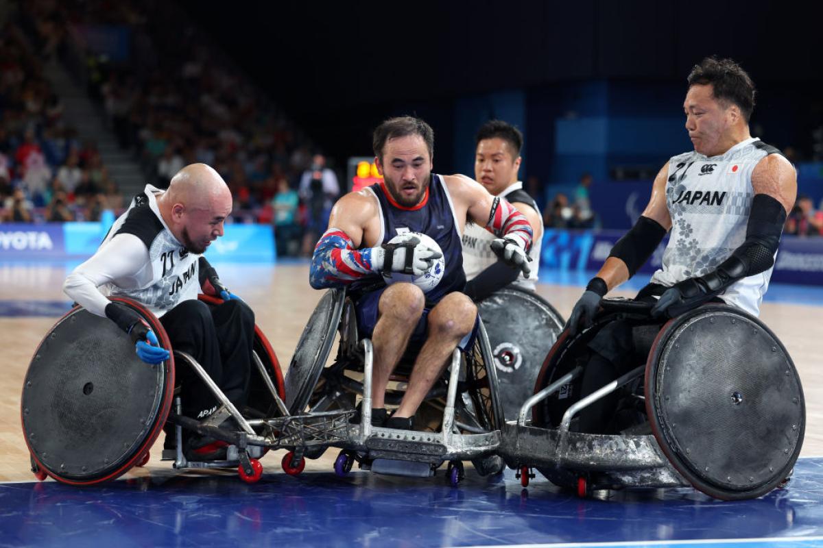 A male wheelchair rugby player carries the ball, while he is surrounded by two players