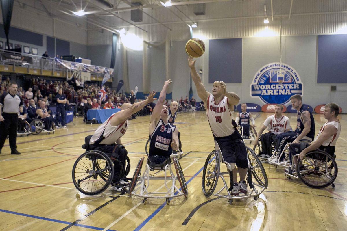 A picture of a man in a wheelchair shooting a Basketball ball between 2 other men in a wheelchair.