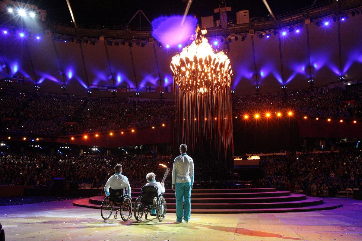 A picture of one man and woman in wheelchairs and one man standing watching a lightened cauldron during a ceremony