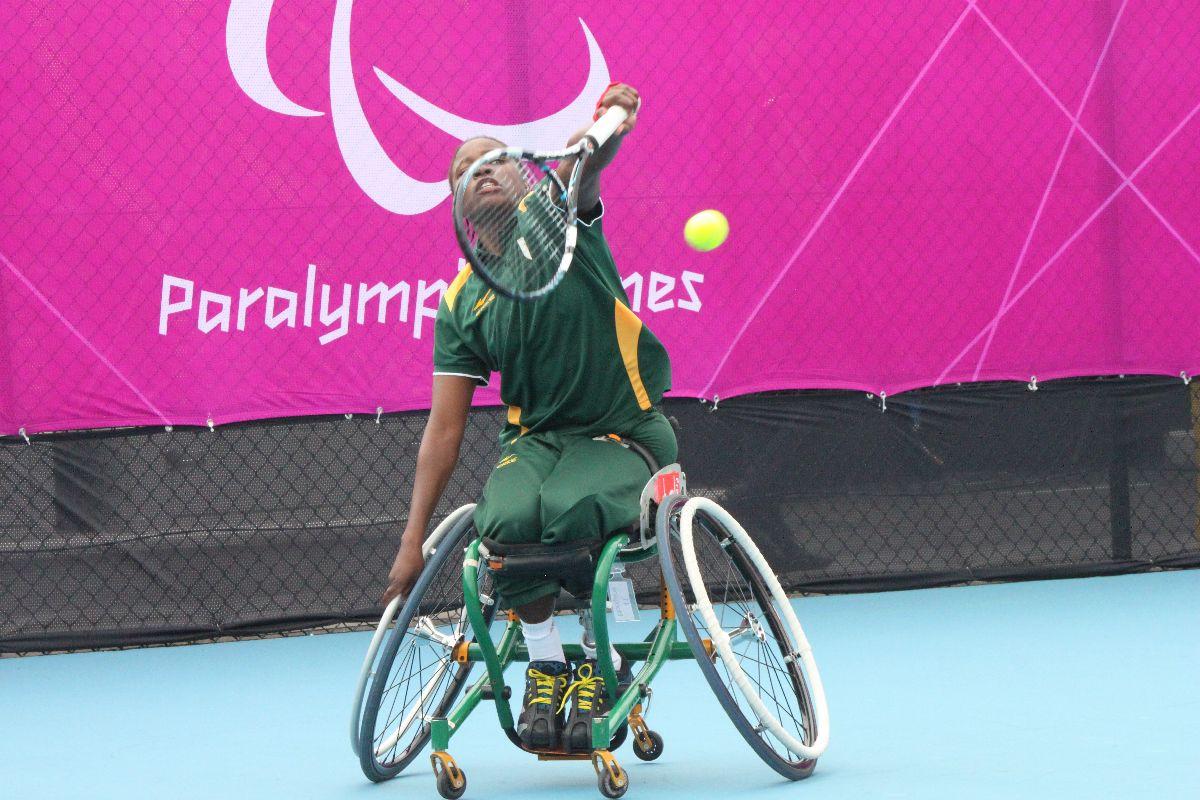 A picture of a black woman in a wheelchair serving during a wheelchair tennis match.