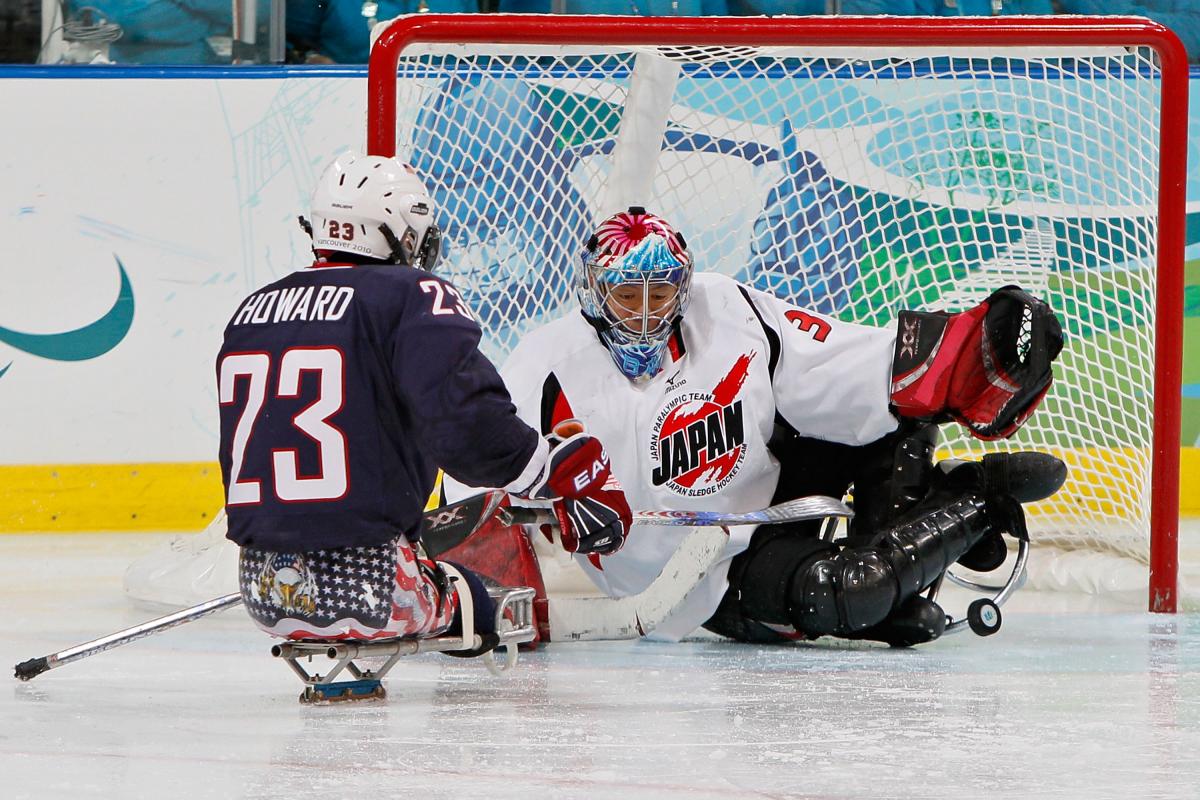 A picture of a man in sledge shooting the puck against the goalkeeper in an ice hockey match