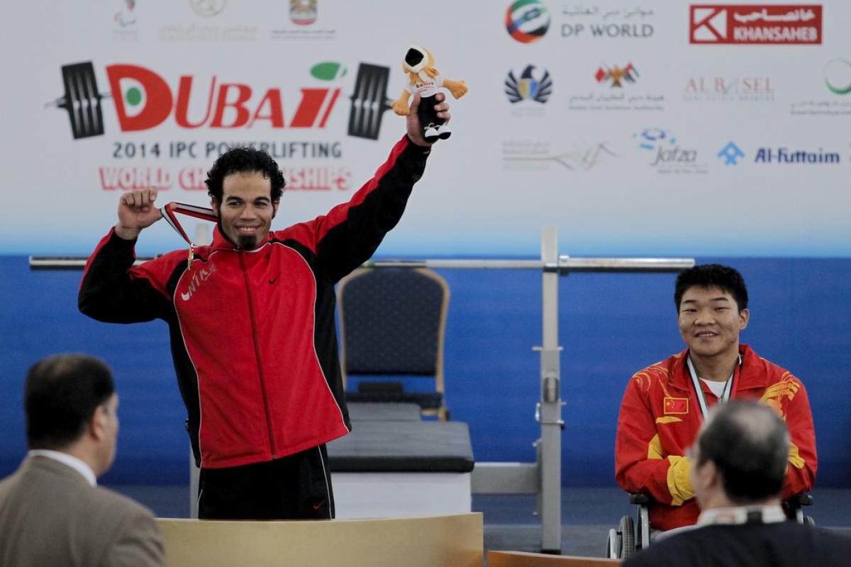 An athlete shows off his medal to a crowd, holding up a stuffed animal mascot with the other hand. 