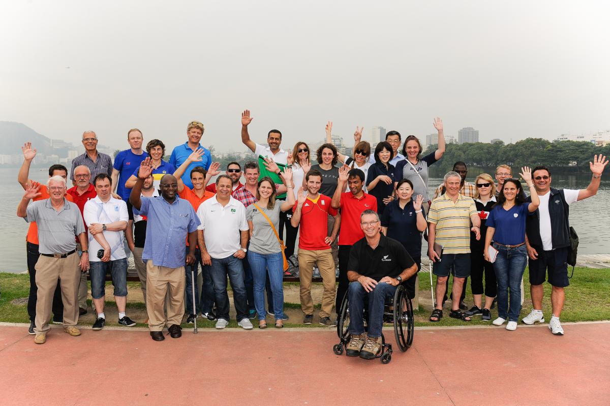 A group of people pose for a photo, waving in front of a body of water.