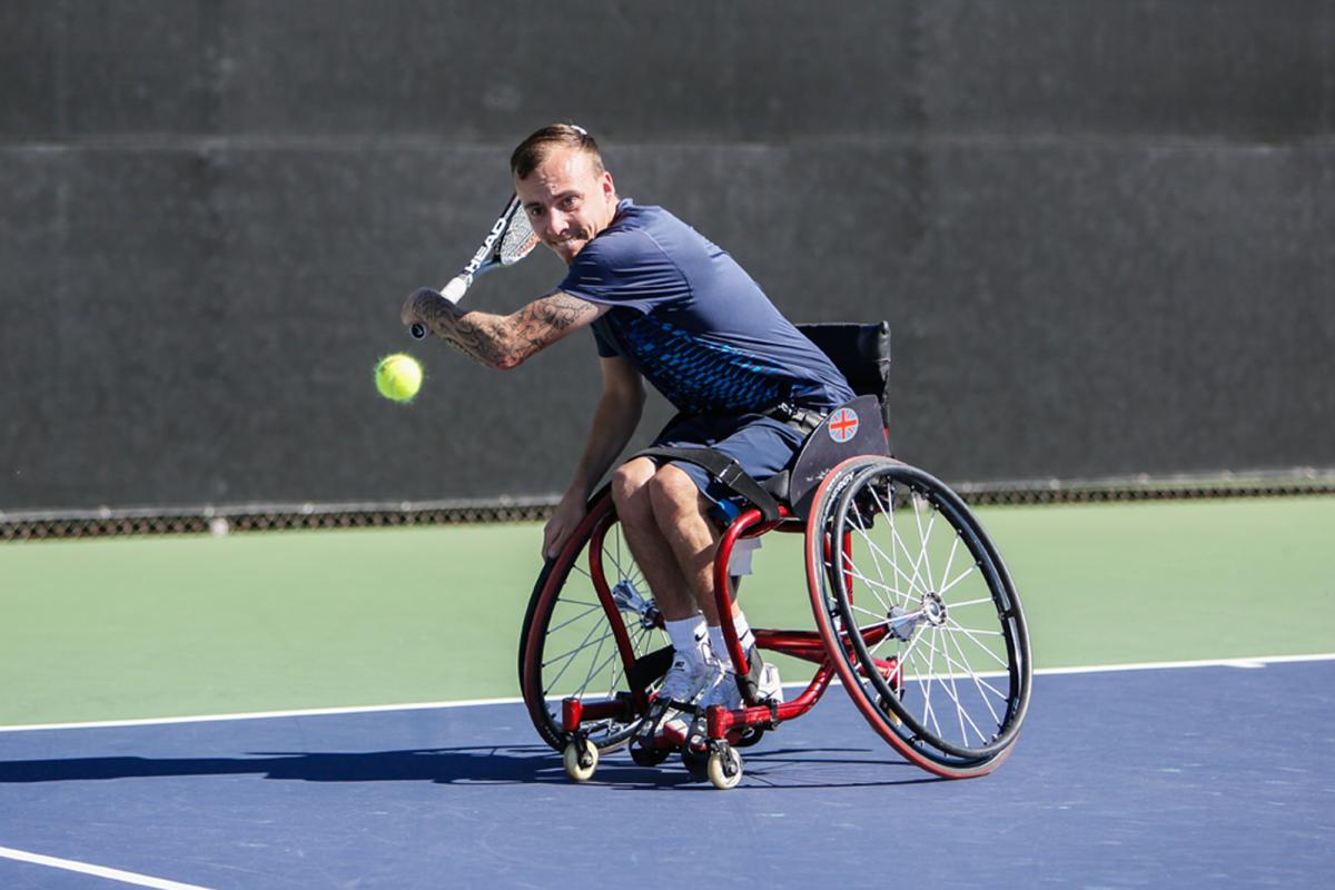 A man in a wheelchair swings back a tennis racket in preparation of hitting a backhand.
