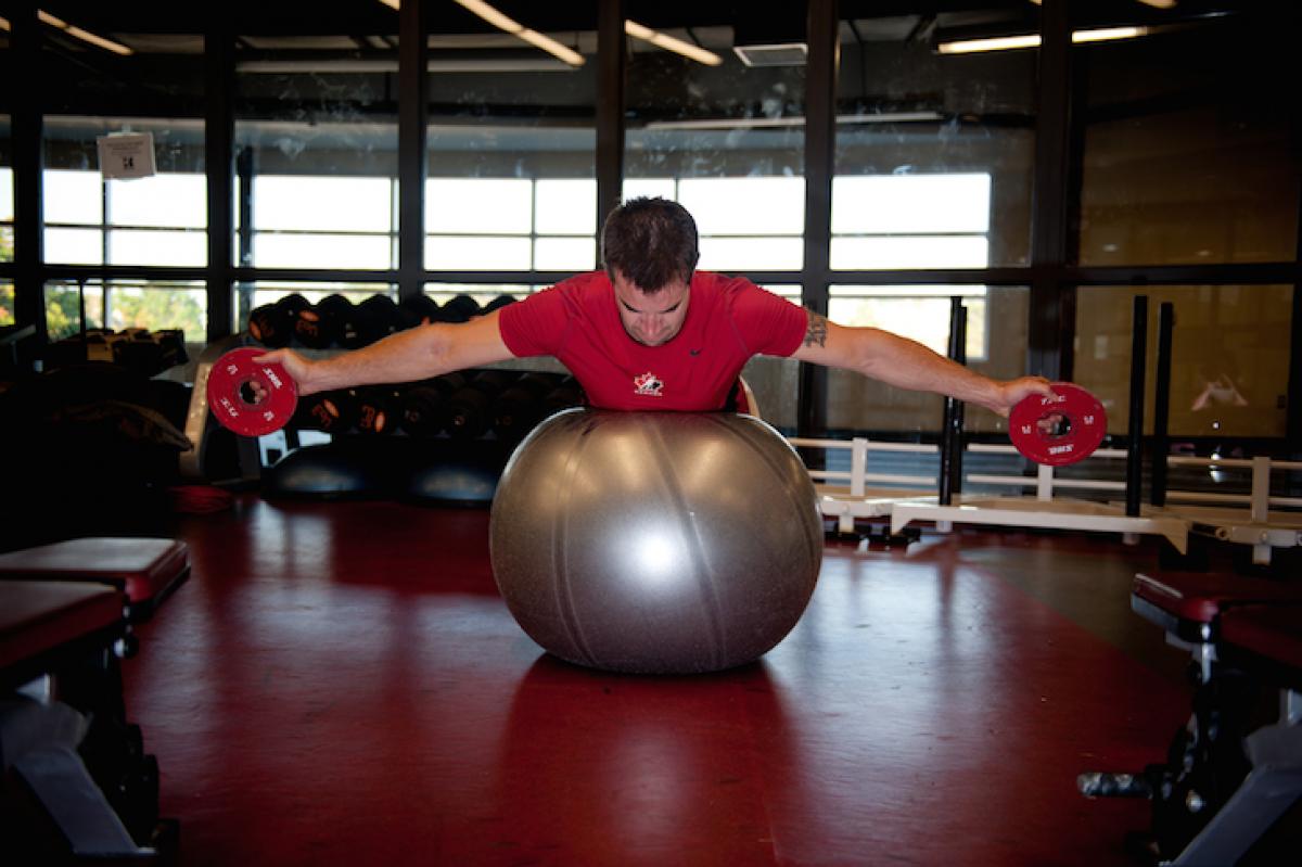 Kevin Rempel An athlete lies on his stomach on a stability ball with both his arms stretched out to the sides holding weights.
