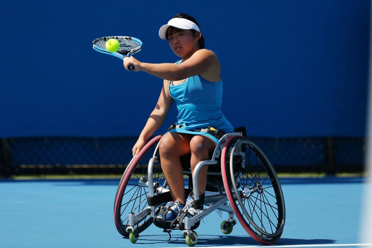 A woman in a wheelchair hits a tennis ball as a backhand.