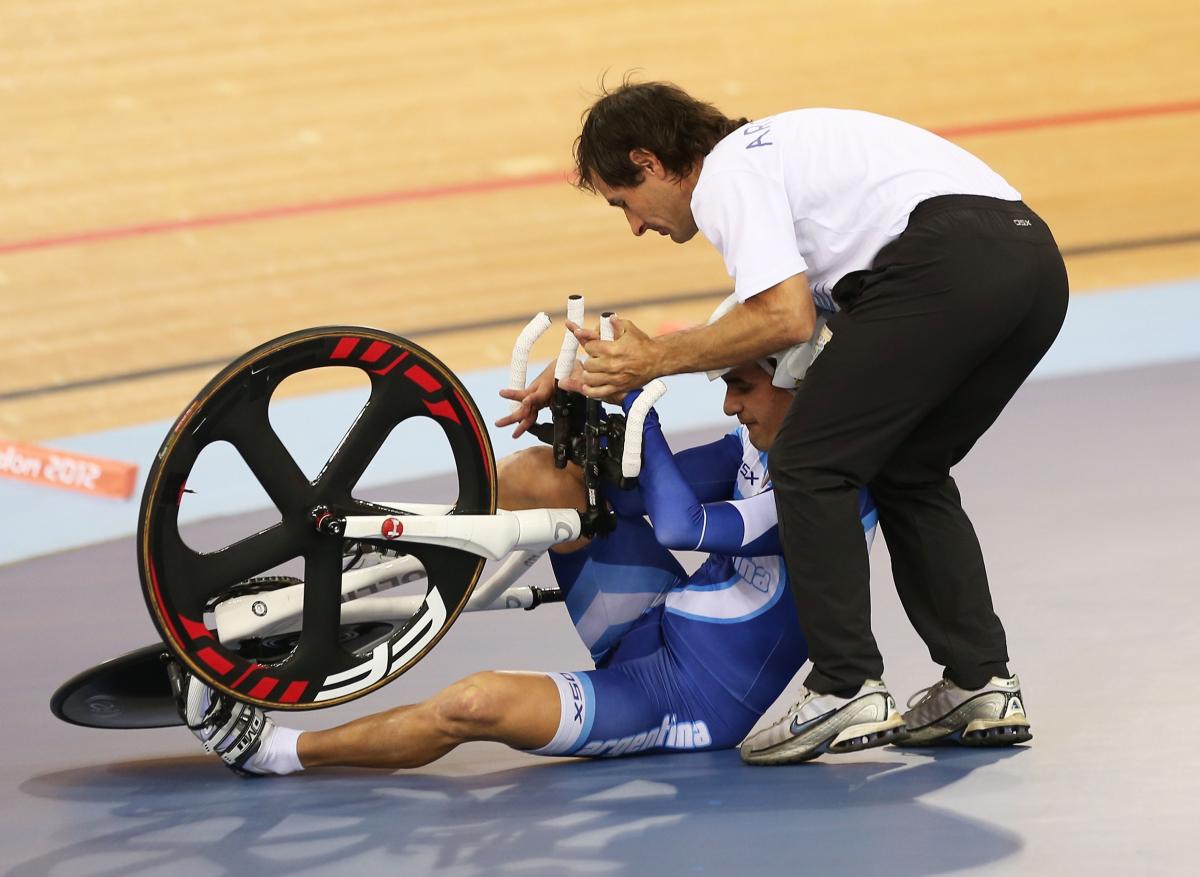 A man sits beside the track trying to get up with his bike with another man assisting him.