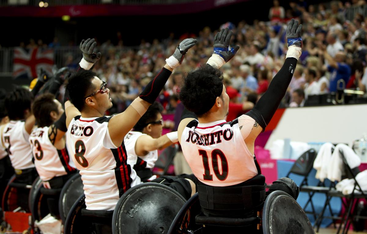 Wheelchair rugby players shown from behind raise their arms and wave to a crowd.