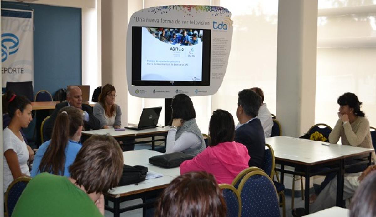 People sitting on chairs around tables, looking at a screen. Classroom atmosphere