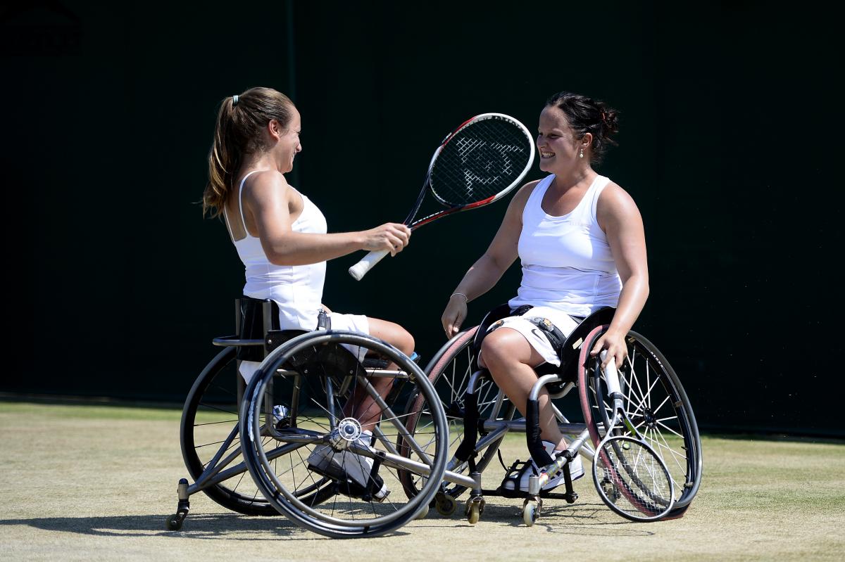 Two women in wheelchairs look at each other smiling on a grass tennis court.