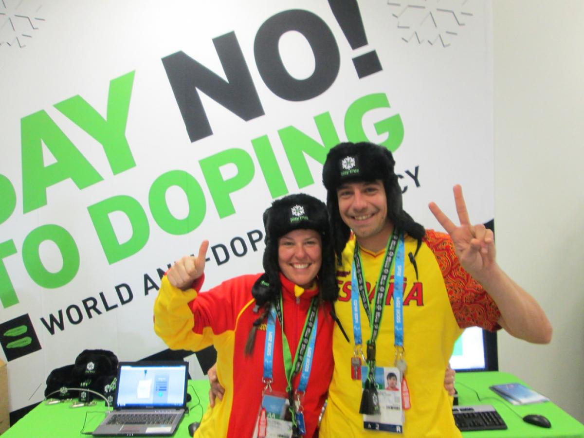 Two people give a thumbs up and a peace sign in front of a "Say No! to Doping" sign.