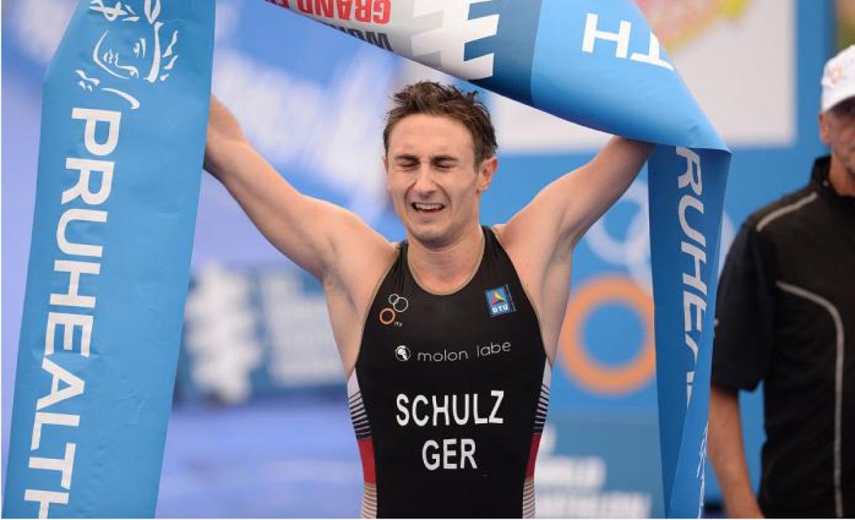 A para-triathlete closes his eyes as he crosses the finish line and raises the finish line banner up in the air.