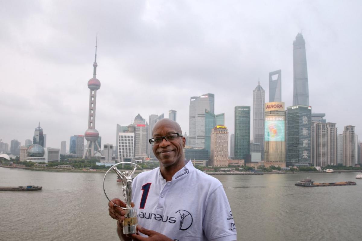 Laureus Chairman Edwin Moses A man's upper body in front of a skyline with skyscrapers in the background. He is holding a silver statue to the camera, smiling